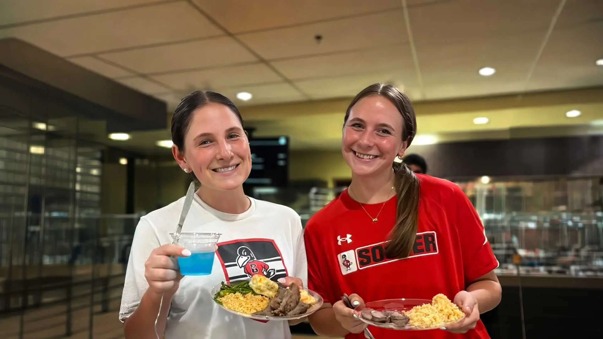 Two students holding plates of food while smiling in middle of dining hall line