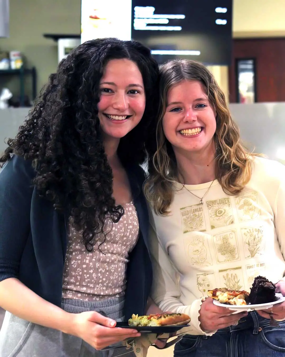Two students smiling in dining hall while holding plates of food
